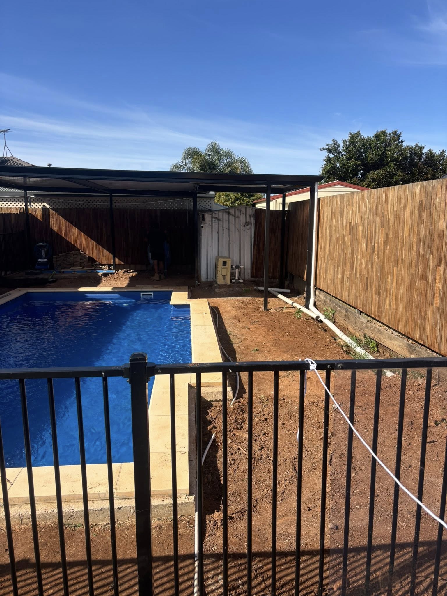 Pool area mid-construction — bare dirt surrounds under a new pergola before paving — May Paving