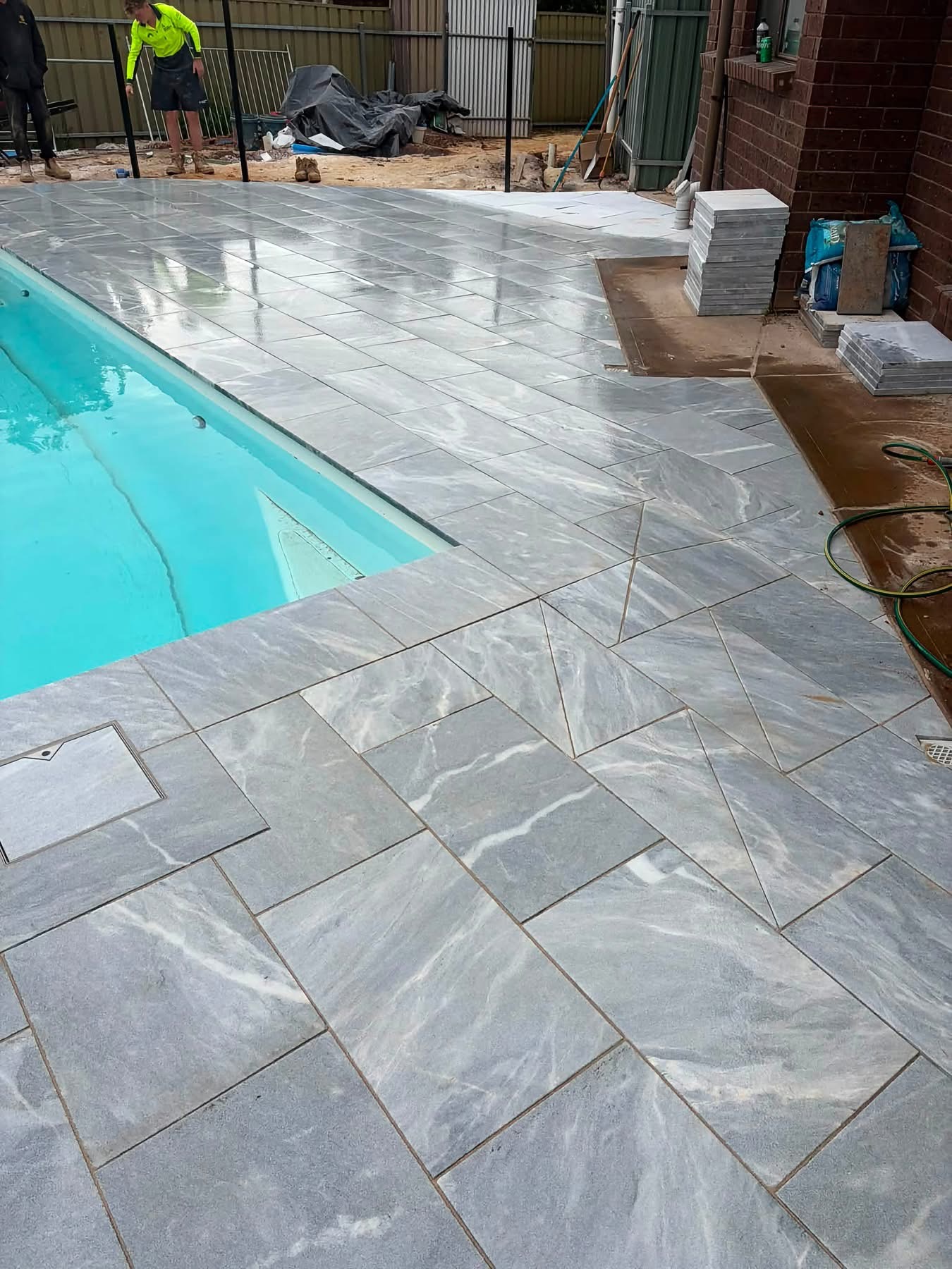 Wet marble-look pavers glistening at the pool edge while the May Paving crew works on the coping