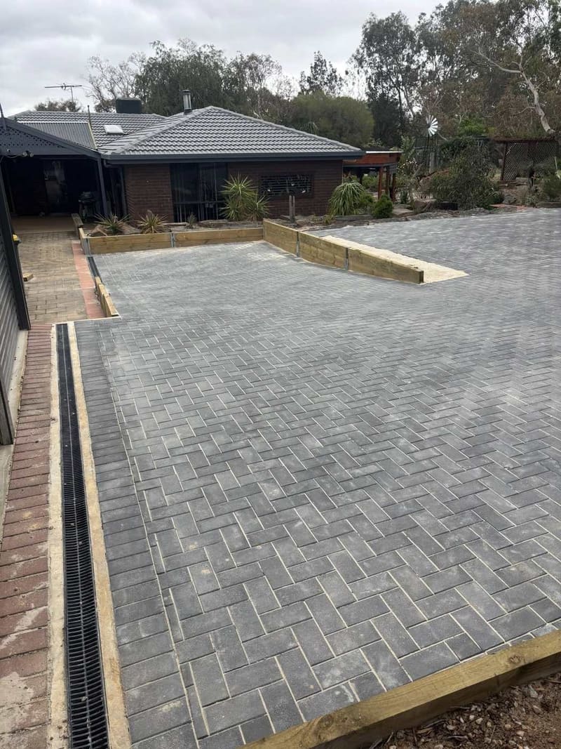 Grey herringbone brick driveway with a grated channel drain along the edge and block retaining walls